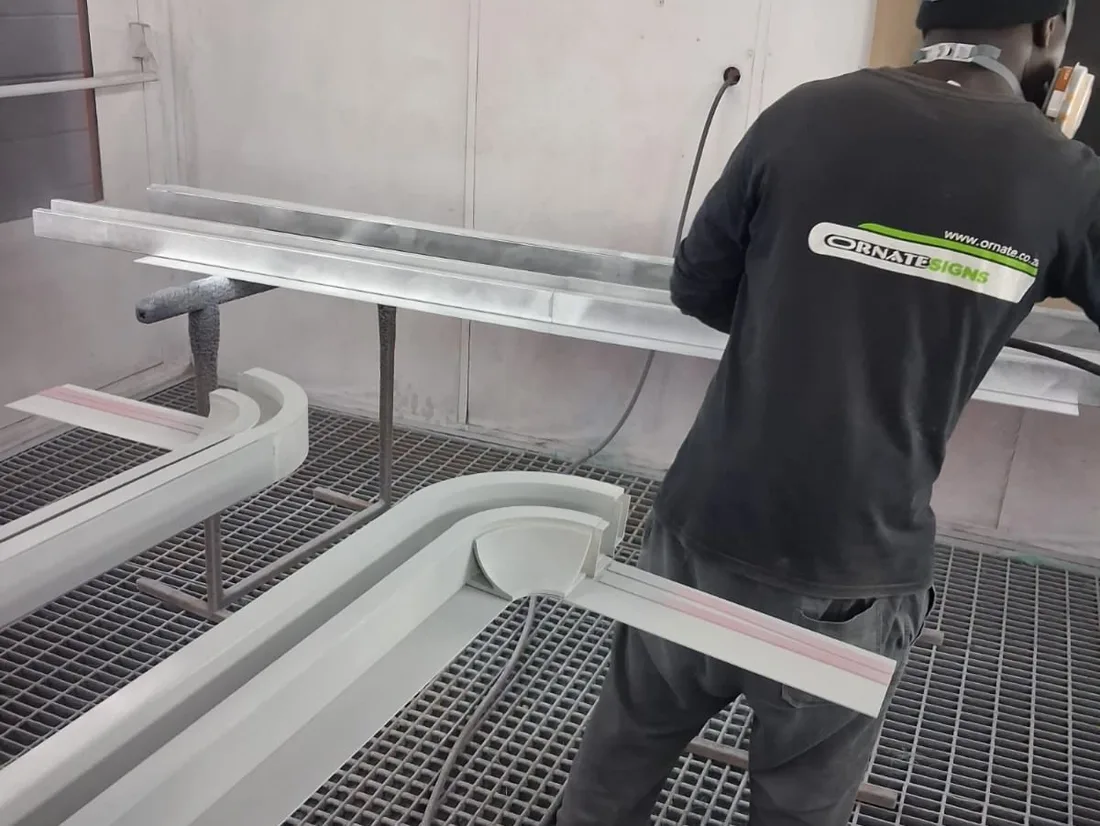 Ornate Signs spraying the assembled ceiling light frame in the booth, with the printed corner section visible in the foreground already coated