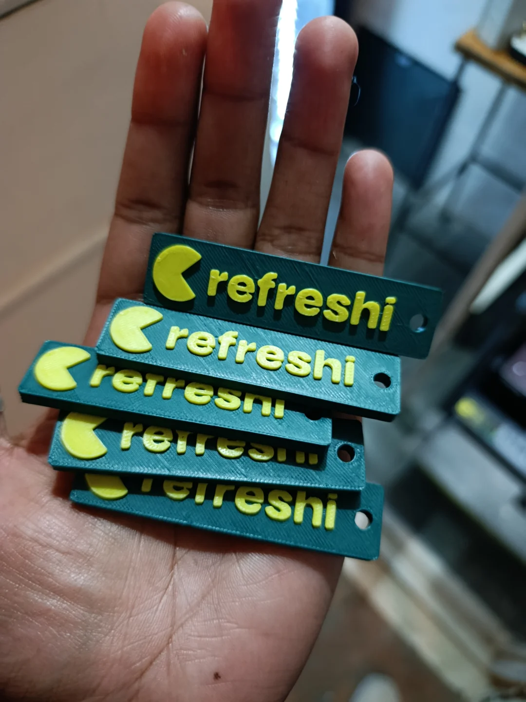 A hand holding five freshly printed Refreshi branded keychains in a stack — teal body with yellow logo and text